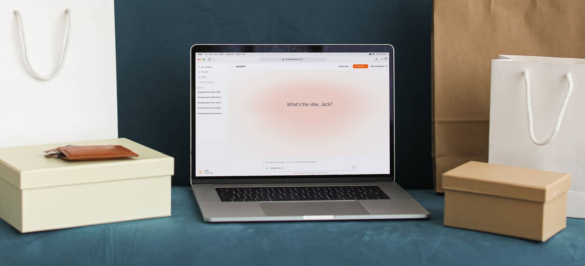 A laptop among shopping bags and gift boxes, open on QioskGPT at qioskmarketplace.com, asking “What's the vibe, Jack?” above a box to describe a campaign